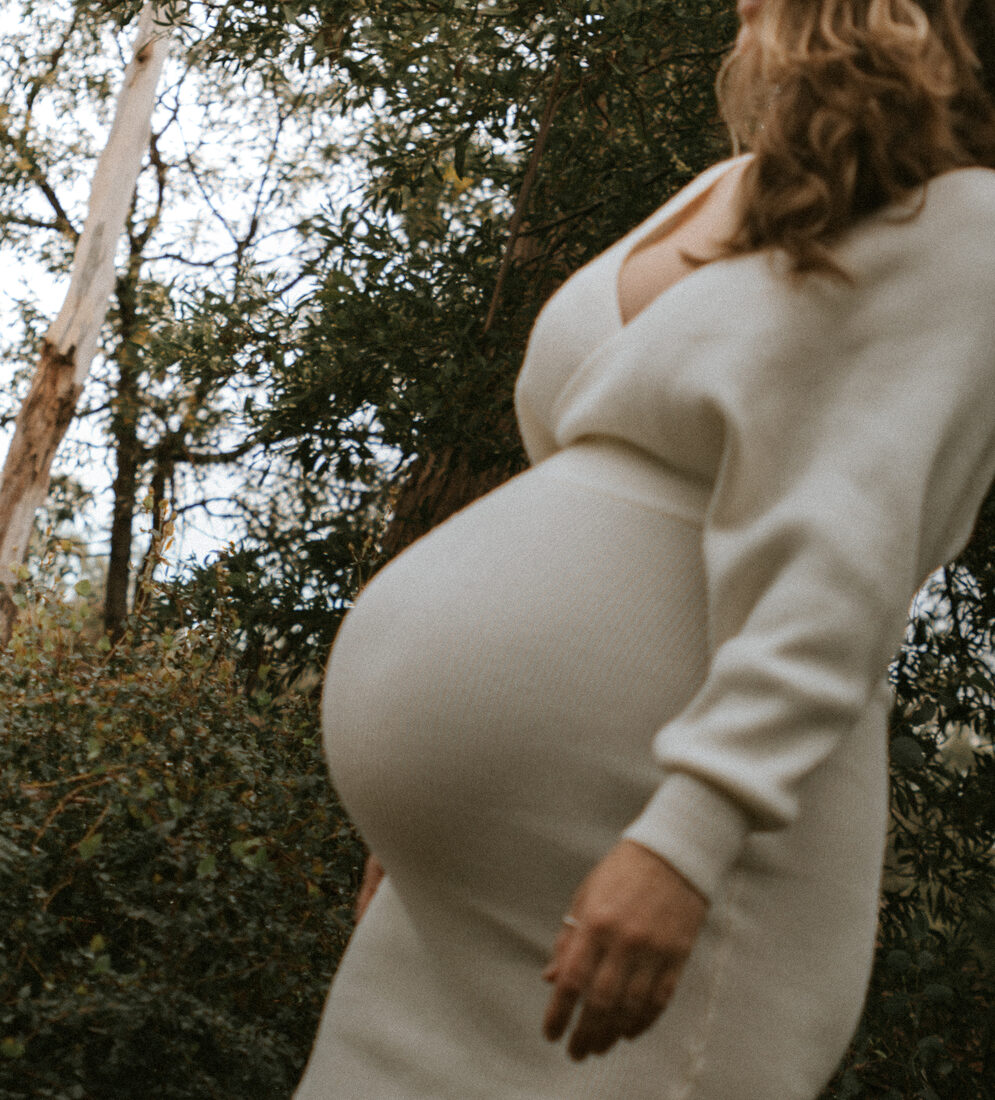 A pregnant woman in a cream dress standing among Australian bushland